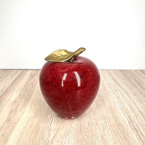 May include: A red decorative apple with a gold-colored leaf. The apple has a glossy finish and sits on a wooden surface. This decorative item is a fruit-themed accent piece.