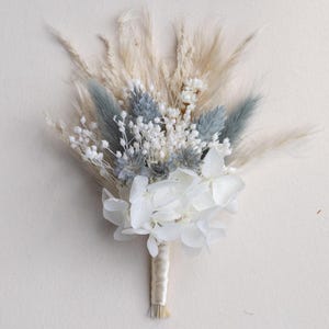 Op de afbeelding: Een klein boeket van gedroogde bloemen met witte hortensia's, blauwe distel en veervormig beige pampasgras. De stelen zijn gebonden met een crèmekleurig lint. Geschikt voor bruiloften of andere speciale evenementen.
