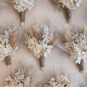 Op de afbeelding: Negen kleine boeketten gedroogde bloemen in tinten van wit en beige. Elk boeket is vastgebonden met touw en bevat gedroogde hortensia's, pampasgras en andere gedroogde bloemen.