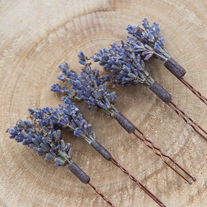 Pode incluir: Quatro grampos de cabelo de lavanda com flores de lavanda secas presas &agrave;s pontas. Os grampos est&atilde;o dispostos em uma fileira sobre uma superf&iacute;cie de madeira.
