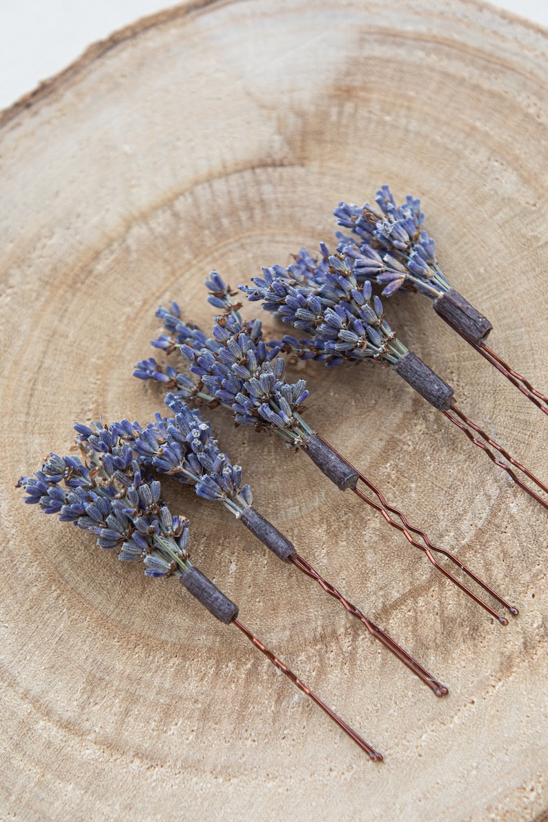 Pode incluir: Quatro grampos de cabelo de lavanda com flores de lavanda secas presas &agrave;s pontas. Os grampos est&atilde;o dispostos em uma fileira sobre uma superf&iacute;cie de madeira.