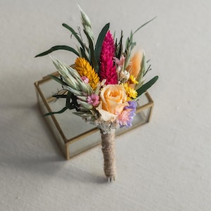May include: A small bouquet of dried flowers in a glass box. The bouquet includes a peach rose, pink and yellow flowers, and dried grasses.