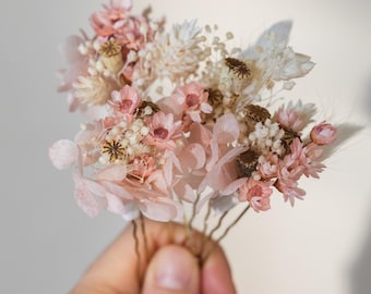 May include: A set of six hair pins adorned with dried pink and white flowers. The flowers are arranged in a cluster and are held together by the pins. The pins are gold in color and have a simple design.