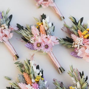 Puede incluir: Una colección de ramos de flores en colores pastel. Cada ramo presenta una variedad de flores secas en tonos rosa, morado, amarillo y verde. Los tallos están envueltos en una cinta rosa claro, creando una estética delicada y elegante.