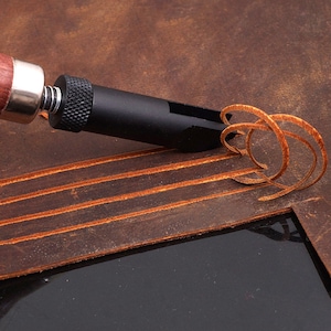 Puede incluir: Una herramienta de trabajo de cuero con un mango negro y una cuchilla afilada. La herramienta se utiliza para cortar una tira de cuero, creando un patrón decorativo.