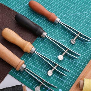 May include: Four leather stitching tools with wooden handles and metal clamps. The tools are arranged on a green cutting mat.