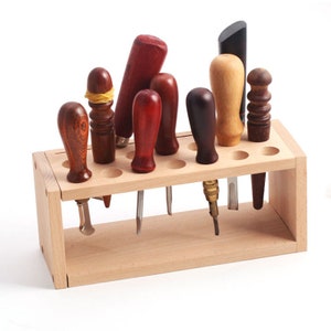 May include: A wooden stand with 10 holes for storing leather working tools. The stand is made of light wood and has a natural finish. The tools are made of different materials, including wood, metal, and plastic.
