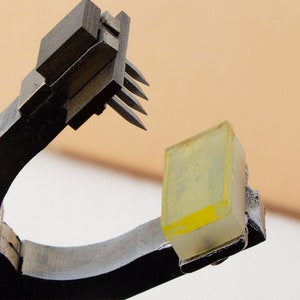 May include: A black metal tool with a set of sharp metal prongs, holding a clear yellow rectangular object.