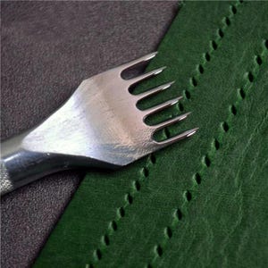 May include: A silver-colored metal leather stitching chisel with six prongs positioned over a piece of dark green leather. The tool features a textured handle and sharp, pointed tines that have created a row of evenly spaced, small, oval-shaped holes.