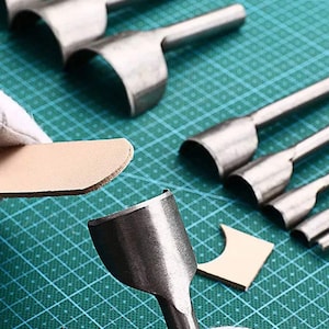 May include: A set of leather craft tools with different sized punches for creating holes in leather. The tools are made of metal and have wooden handles. The tools are laid out on a green cutting mat.