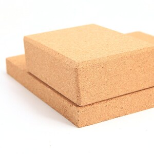 May include: Two rectangular yoga blocks made of light brown cork are stacked on a white background. The top block is slightly smaller than the bottom one. The blocks have a natural, textured appearance, suggesting a focus on eco-friendly materials.