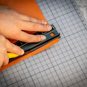 May include: A person using a black metal ruler and a yellow utility knife to cut a piece of orange paper on a gray grid cutting mat.