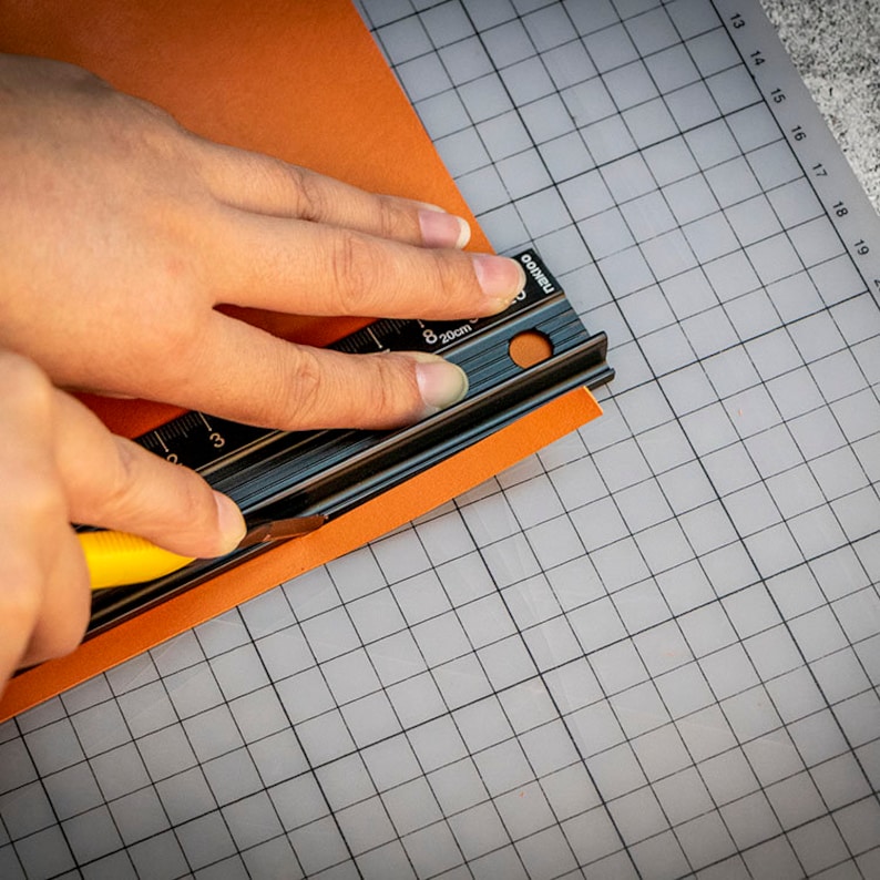 May include: A person using a black metal ruler and a yellow utility knife to cut a piece of orange paper on a gray grid cutting mat.