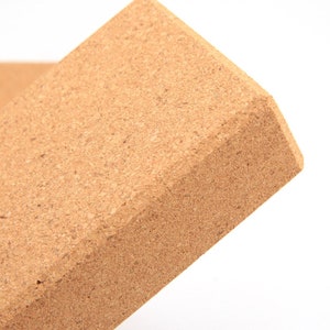 May include: A rectangular yoga block made of natural cork. The block has a light brown color and a textured surface. The edges are beveled, and the block is designed for use in yoga and Pilates.