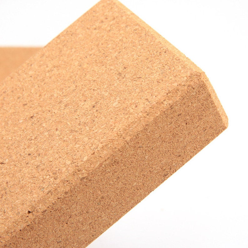 May include: A rectangular yoga block made of natural cork. The block has a light brown color and a textured surface. The edges are beveled, and the block is designed for use in yoga and Pilates.