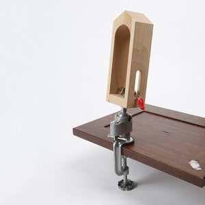 May include: A wooden clamp with a red handle, attached to a silver clamp, is mounted on a brown wooden surface.