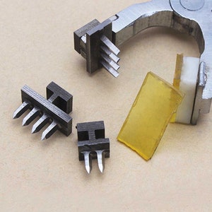 May include: Four metal tools with sharp points, used for punching holes in leather or other materials. The tools are arranged on a light brown surface. One tool is attached to a metal clamp.