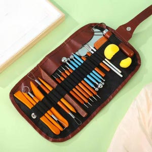 May include: A brown faux leather roll-up case containing a set of sculpting tools. The set includes various metal loop tools, wooden-handled needles, ball styluses with blue handles, orange plastic scrapers, and two yellow sponge pieces.