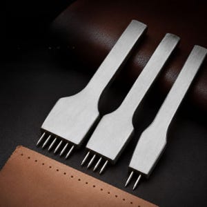 May include: Three silver-toned metal leather stitching chisels with varying prong counts, including a six-prong, a four-prong, and a two-prong tool, positioned above a piece of tan leather with a pre-punched row of holes along the edge.