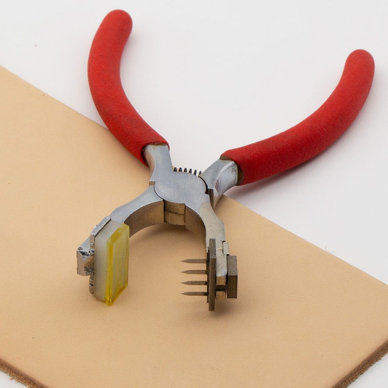 May include: A red-handled leather punch tool with a metal head and a yellow plastic piece. The tool is lying on a piece of tan leather.