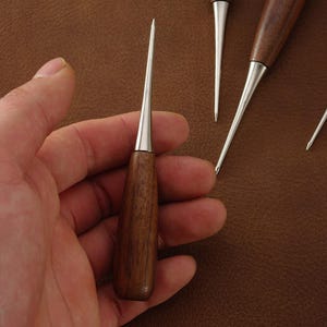 May include: A set of leatherworking awls featuring polished silver-toned metal tips and smooth, dark brown wooden handles. The tools have a tapered, sharp point design, with one awl held upright and others positioned nearby on a textured surface.