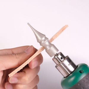 May include: A silver-colored metal burnishing tool with a pointed, tapered tip attached to a power drill chuck. The tool is positioned against a thin, rectangular strip of light tan leather, showing the tool's textured, abrasive surface.