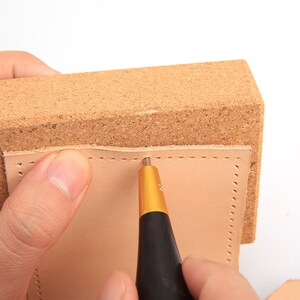 May include: A close-up of leather crafting. A light brown piece of leather is being worked on with a black and gold tool. The leather is placed on a brown cork block. The tool is creating a line of small holes.