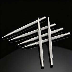 May include: A set of six silver-colored metal stylus tools with pointed tips and cylindrical bodies. Each tool features a textured, cross-hatched knurled grip section in the center, while the ends are smooth and polished, reflecting light against a dark background.