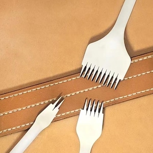 Op de afbeelding: Drie zilverkleurige metalen stikbeitels voor leer met variërende aantallen tanden, waaronder een met twee tanden en twee met meerdere tanden, geplaatst bij een beige leren strook met twee parallelle rijen witte stikseldetails.