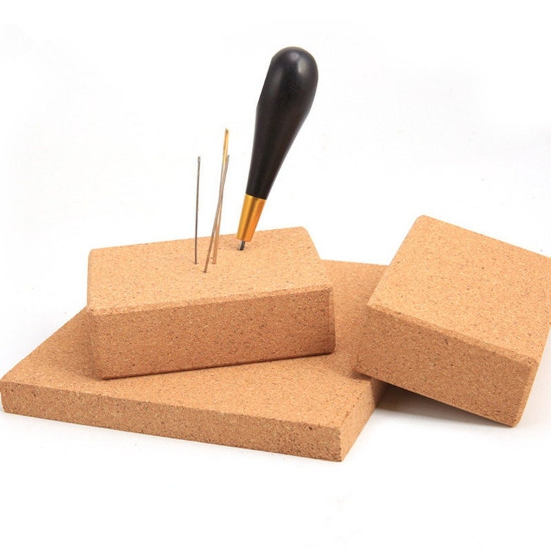May include: A set of cork blocks in various sizes, with a black-handled awl and several needles inserted into one block. The cork has a light brown color, and the awl has a gold ferrule. The needles are silver.