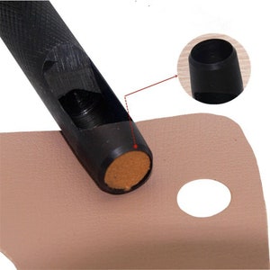 May include: A black leather punch tool with a brown cushioned base, used to create holes in leather. The tool is shown punching a hole in a piece of light brown leather.