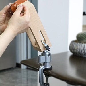 May include: A wooden leather working tool with a clamp base and a swivel arm. The tool is used to hold leather while stitching. A person is using the tool to stitch a piece of brown leather.