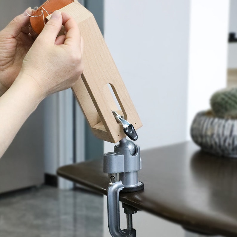 May include: A wooden leather working tool with a clamp base and a swivel arm. The tool is used to hold leather while stitching. A person is using the tool to stitch a piece of brown leather.