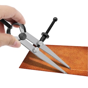 May include: A silver and black leather crafting tool with a pointed tip and a knob for adjusting the width. The tool is being used to draw a line on a piece of brown leather.