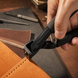 May include: A black metal leather stitching groover tool with a spring mechanism and black foam-covered handles, used to create a line of evenly spaced holes along the edge of a piece of tan leather, with other leather crafting tools visible nearby.