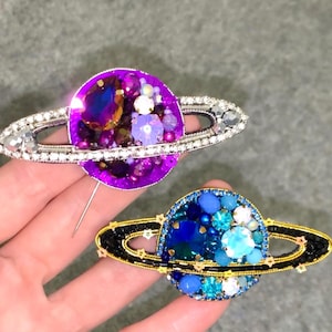 Puede incluir: Dos broches con forma de planeta que presentan un intrincado trabajo de cuentas y cristales. El broche superior es morado con anillos plateados, mientras que el inferior es azul con anillos negros. Ambas piezas tienen piedras facetadas.