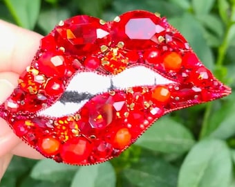 May include: A red lips brooch with white lips and a beaded design. The brooch is made of red and orange beads and gems. The text "@aisha_fashion_accessories" is at the bottom of the image.