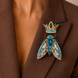 May include: A decorative brooch shaped like a fly with a crown positioned above it. The insect features bright blue, gold, and clear crystals, with intricate beadwork detailing its wings and body. The crown is accented with blue and gold crystals.