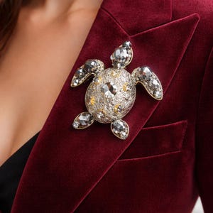 Puede incluir: Un broche de tortuga enjoyado con cristales transparentes y amarillos está prendido a una blazer de terciopelo burdeos. El broche presenta un gran cristal central y cristales más pequeños en la cabeza, aletas y cola. Accesorio decorativo.