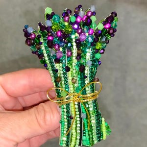 May include: A beaded brooch shaped like a bouquet of flowers, featuring vibrant green, purple, and dark blue beads. The stems are wrapped in green wire and tied with a thin gold ribbon. The text AishaAccessoriesKZ is visible at the bottom.