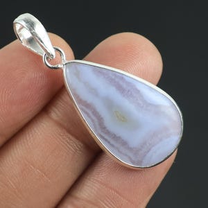 Puede incluir: Un colgante en forma de lágrima con una piedra lisa de bandas de color violeta claro y blanco, engastada en un marco de metal plateado pulido con una anilla plateada a juego en la parte superior para sujetarlo a una cadena o cordón.