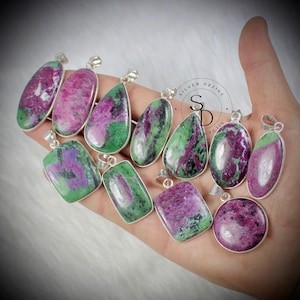 Puede incluir: Una colección de 12 colgantes de piedras preciosas verdes y rosas con engastes de plata. Los colgantes son de varias formas y tamaños, incluyendo ovalados, en forma de lágrima y rectangulares.