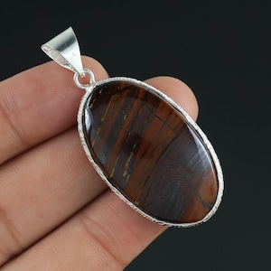 Puede incluir: Colgante ovalado con una piedra preciosa de ojo de tigre pulida, que muestra bandas marrones y doradas. La piedra está engastada en un marco de metal plateado con un borde decorativo y un lazo para una cadena.