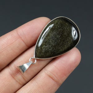 May include: A teardrop-shaped pendant featuring a polished, dark green gemstone with gold flecks. The stone is set in a silver-colored metal frame and has a small loop and bail for a chain. The pendant is displayed against a dark background.