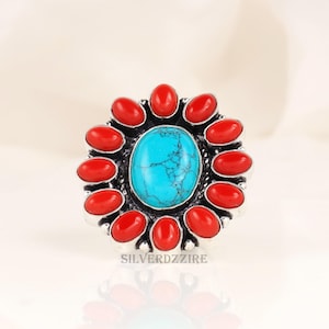 May include: A silver ring with a central oval turquoise stone, encircled by red coral stones. The ring's design resembles a flower, with the turquoise as the centrepiece. The ring is set against a soft, neutral background. The ring is a statement piece.