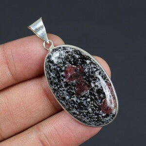 Könnte beinhalten: Ovaler Anhänger mit einem schwarz-weiß gesprenkelten Stein mit roten Einschlüssen, gefasst in einem silberfarbenen Metallrahmen. Der Anhänger hat eine Schlaufe für eine Kette. Der Stein hat ein einzigartiges Muster und eine polierte Oberfläche.