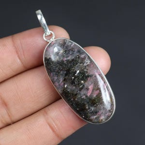 May include: A polished, oval-shaped pendant with a silver-colored metal frame. The pendant features a mix of black, gray, and pink minerals. The pendant is designed to be worn as a necklace.