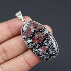 Op de afbeelding: Ovale hanger met een zilverkleurige beugel. De hanger heeft een gespikkeld patroon van zwarte, witte en rode mineralen. De hanger wordt in een hand gehouden.