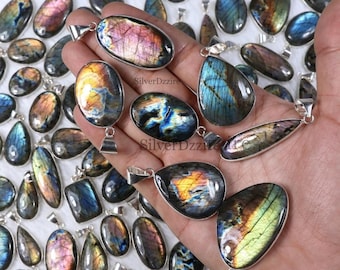 Può includere: Una collezione di ciondoli in labradorite lucida di varie forme, tra cui ovali, a goccia e a pera, caratterizzati da riflessi iridescenti blu, oro e rosa. Ogni pietra è montata su una contromaglia in metallo argentato su sfondo bianco.