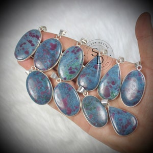 Puede incluir: Una colección de colgantes de piedras preciosas con engaste plateado. Los colgantes presentan una piedra azul-gris con inclusiones rojas y verdes. Los colgantes tienen varias formas ovaladas y de lágrima.